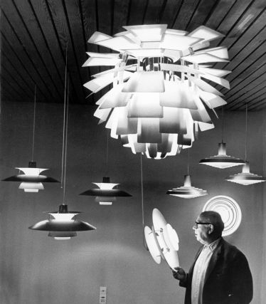 Une photographie historique en noir et blanc de Poul Henningsen avec ses lampes emblématiques pour Louis Poulsen. Au premier plan, Henningsen tient un modèle de lampe dans sa main, tandis que plusieurs de ses créations sont suspendues au-dessus de lui, notamment la célèbre lampe Artichoke, qui diffuse une lumière douce et non éblouissante grâce à ses nombreuses lamelles.