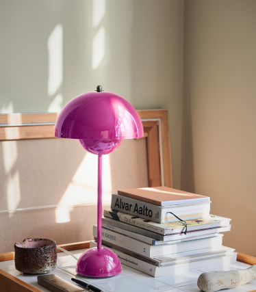 Une lampe de table rose vif avec un abat-jour brillant en forme de dôme trône sur un bureau à côté d'une pile de livres de design, dont certains ouvrages d'Alvar Aalto. Une douce lumière naturelle illumine la scène, mettant en valeur la lampe et projetant de légères ombres sur le fond neutre.