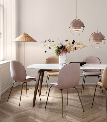 Une salle à manger douce et élégante avec une table en marbre blanc et des chaises rose pastel et beige sur des pieds en laiton. Au-dessus de la table sont suspendues deux suspensions rondes dans des tons beige chaud avec des détails en laiton. Un grand lampadaire avec un abat-jour tissé se trouve près de la fenêtre, et un vase avec des compositions florales séchées décore la table. La pièce est dotée d'un parquet à chevrons clair et d'une palette de couleurs neutres et apaisantes.