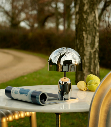 Lampe à poser réfléchissante Tom Dixon sur une table de jardin à côté d'un magazine, entourée d'une chaude lumière du soir dans la verdure.