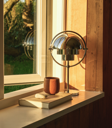 Lampe à poser chromée sur un rebord de fenêtre dans la lumière du soir, à côté d'un livre et d'une tasse en céramique.