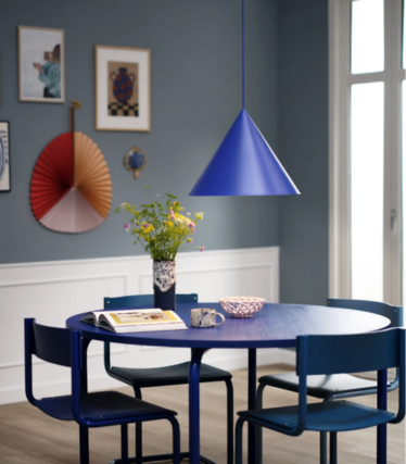 Suspension bleue en forme de cône au-dessus d’une table à manger ronde bleue avec un bouquet de fleurs, dans une salle à manger avec un mur bleu et des œuvres d’art.